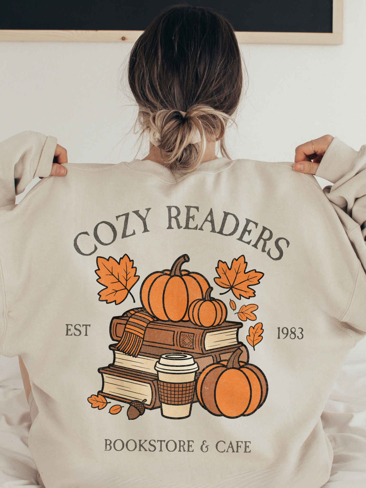 Back view of oversized sand Gildan sweatshirt featuring Cozy Readers Bookstore and Cafe graphic with pumpkins, books, and acorns. Model has hair in a bun and is pulling at her collar.