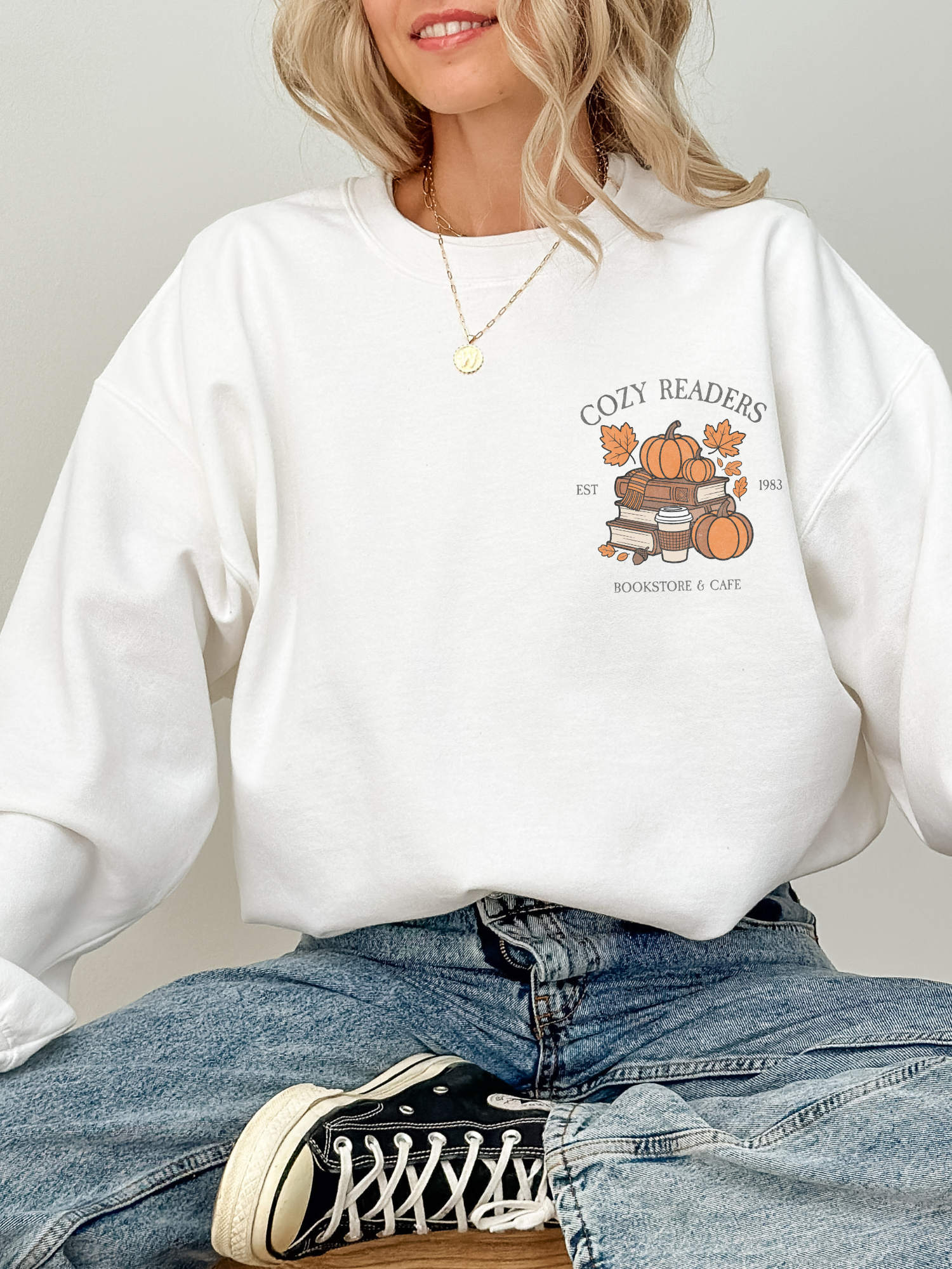 Model sitting cross-legged in white Cozy Readers sweatshirt with chest graphic of pumpkins, books, and coffee cup. Styled casually with light denim and sneakers.