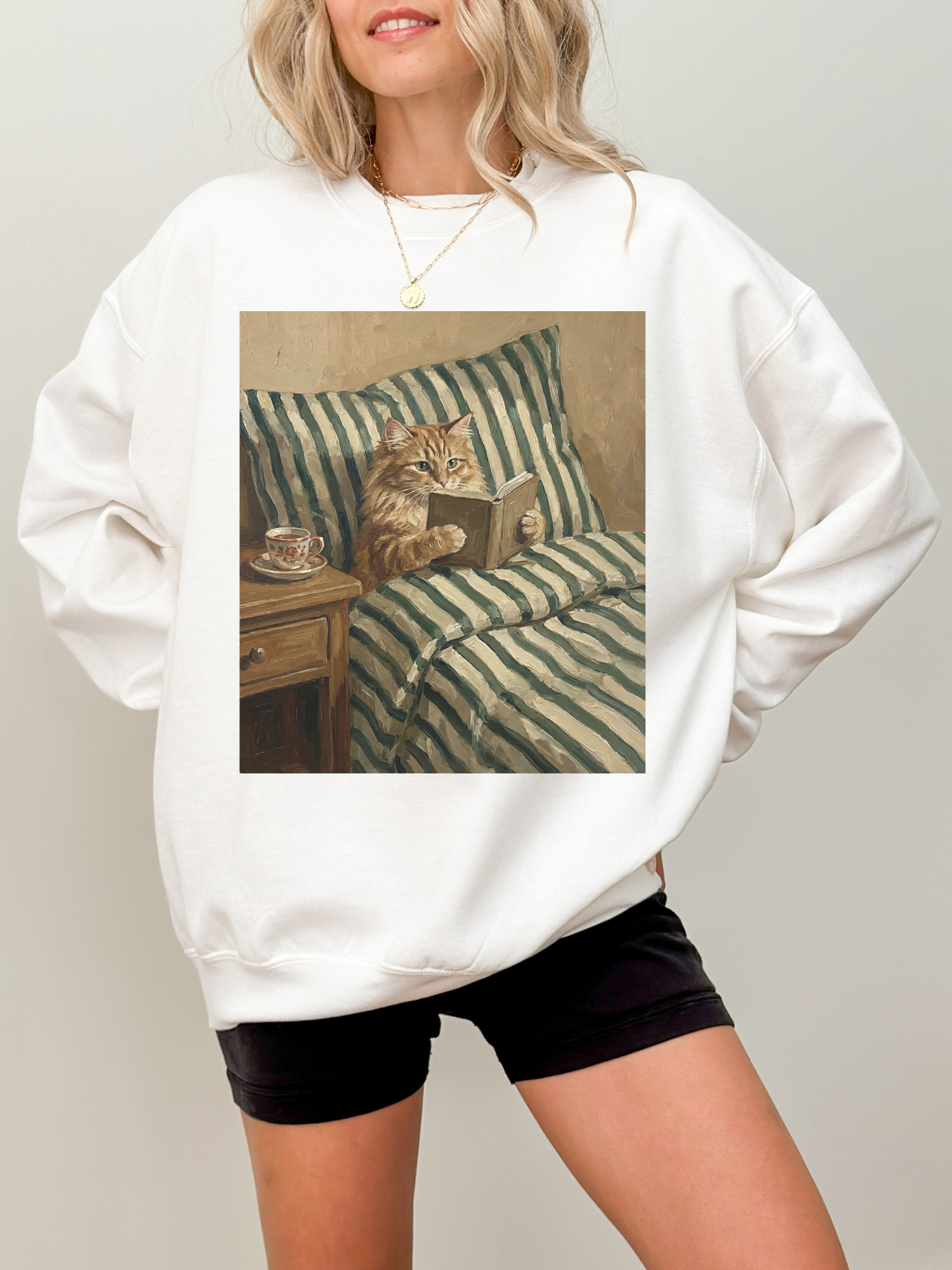 White oversized sweatshirt featuring a vintage style illustration of a cat reading a book in bed with striped bedding and a tea cup on a nightstand. Styled on a model indoors with a minimal background, creating a cozy book lover aesthetic perfect for readers, cat lovers, and fans of relaxing reading vibes.