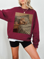 Maroon sweatshirt featuring a cozy vintage style cat reading a book in bed with a bedside table and tea cup, worn by a model indoors. The rich color and relaxed fit create a warm and inviting aesthetic, perfect for book lovers, readers, and anyone who loves cozy cat themed clothing.