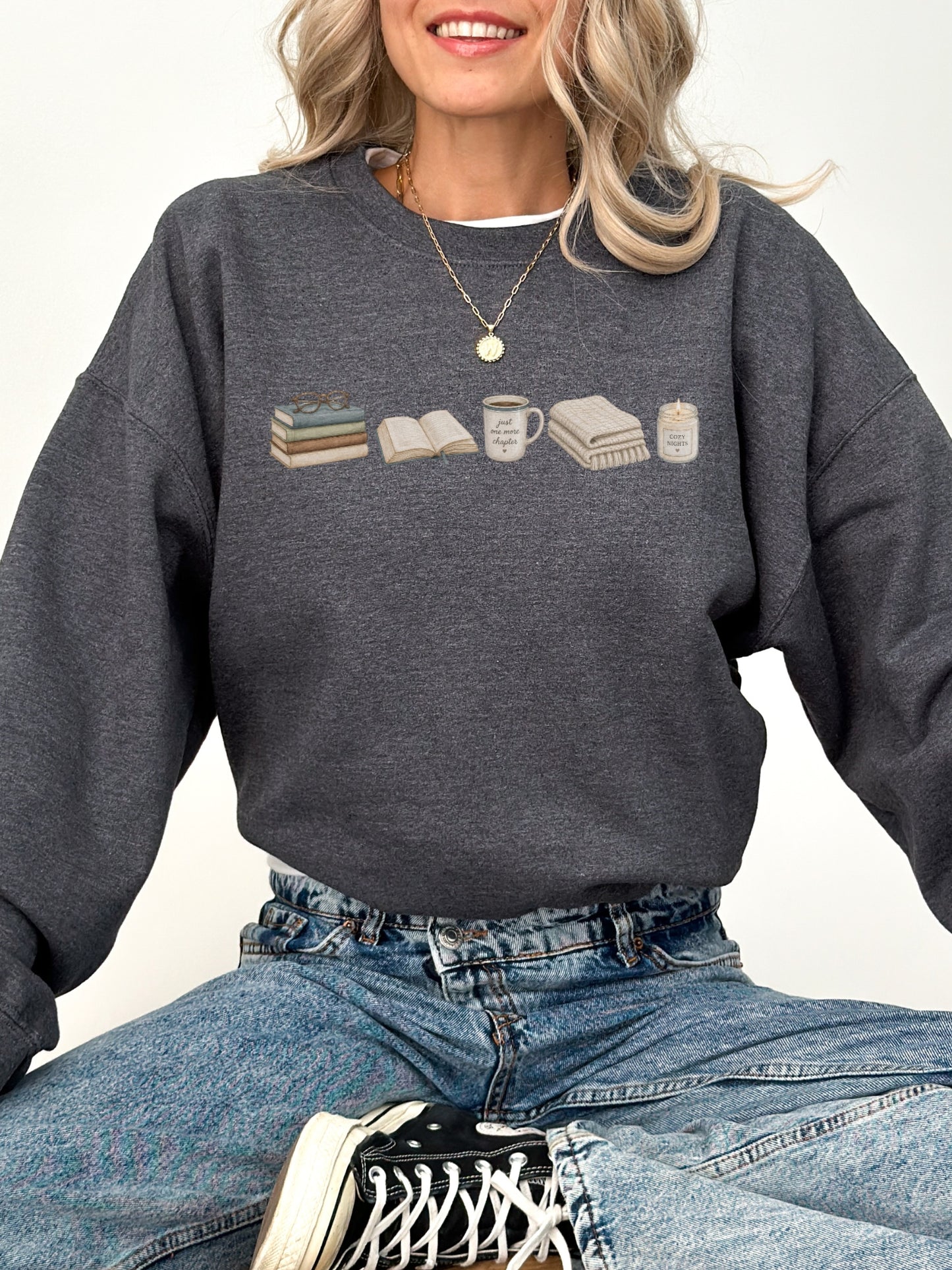 Woman wearing a dark heather Gildan book lover sweatshirt with books, coffee mug, blanket and candle design