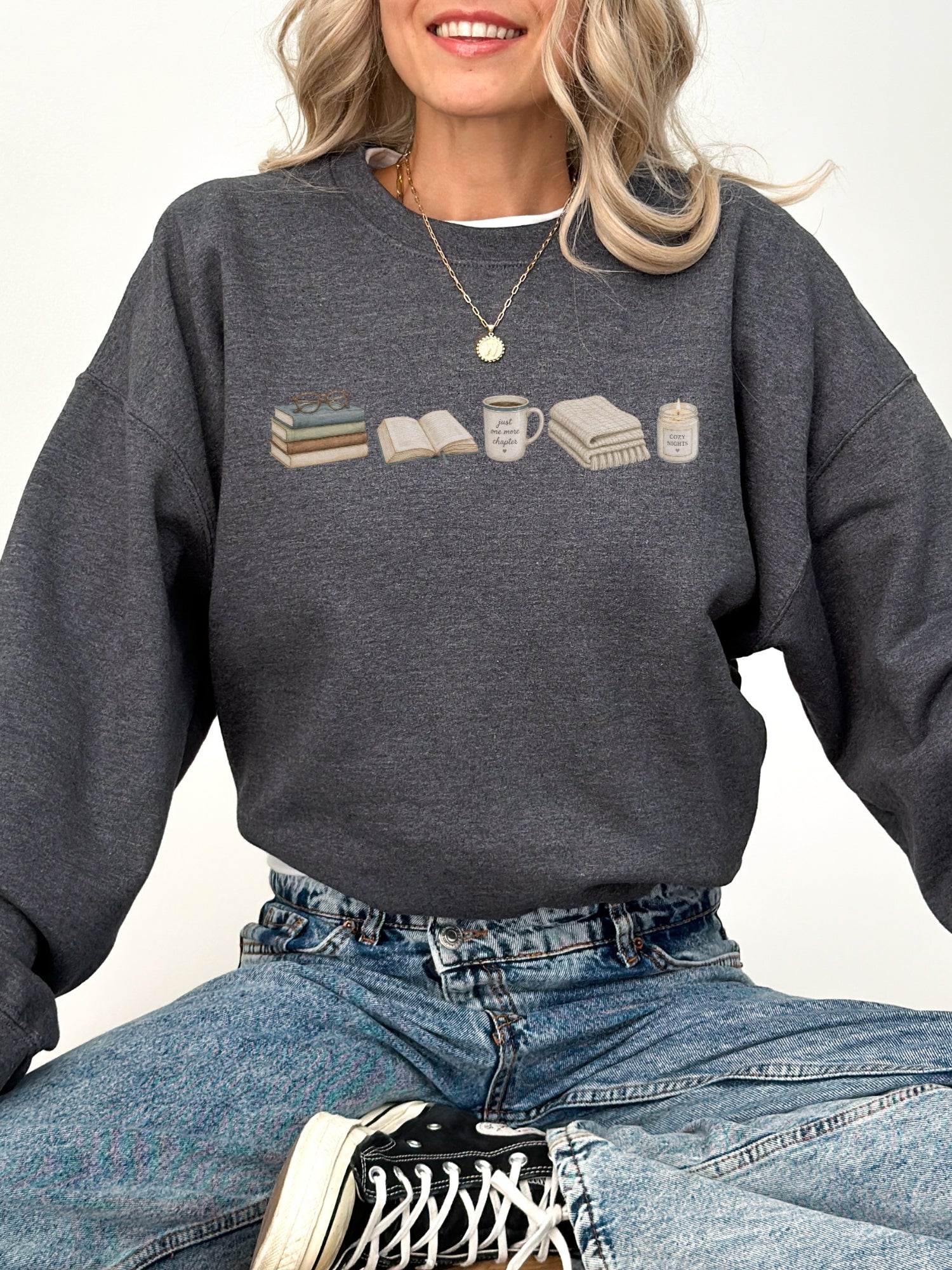Woman wearing a dark heather Gildan book lover sweatshirt with books, coffee mug, blanket and candle design