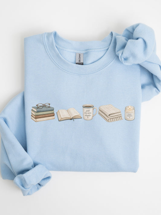 Folded light blue Gildan sweatshirt with books, Just One More Chapter coffee mug, blanket and cozy candle design