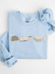 Folded light blue Gildan sweatshirt with books, Just One More Chapter coffee mug, blanket and cozy candle design