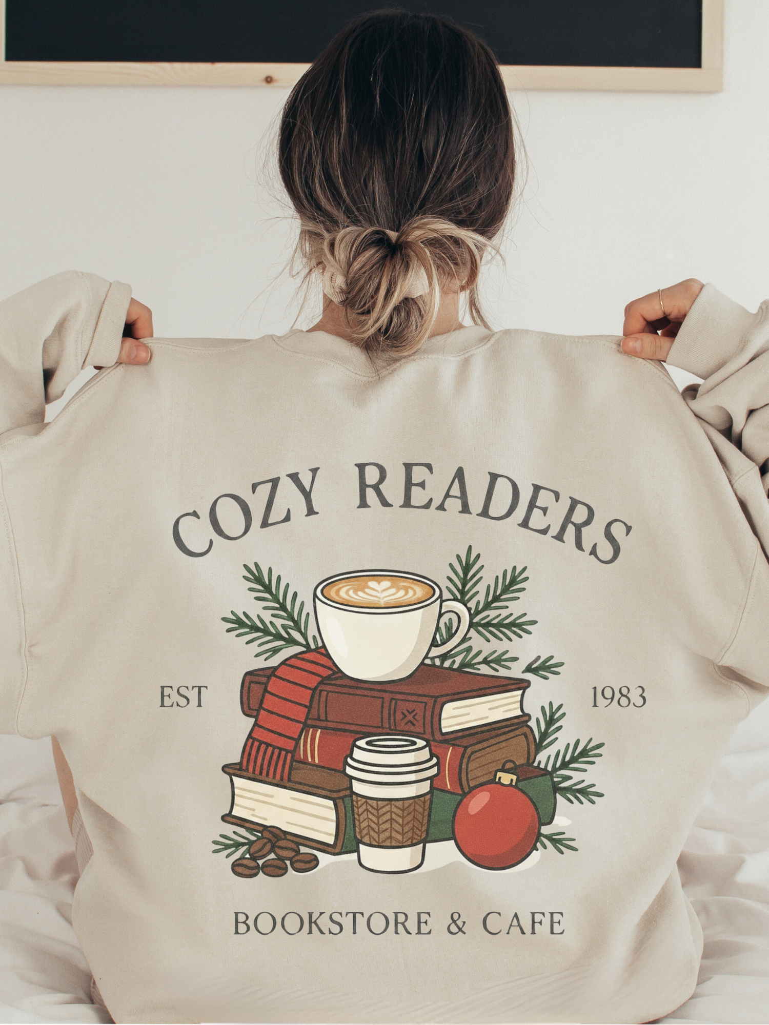 girl holding out the back of a sand sweatshirt showing a cozy reader graphic with books, coffee, and ornaments