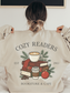 girl holding out the back of a sand sweatshirt showing a cozy reader graphic with books, coffee, and ornaments