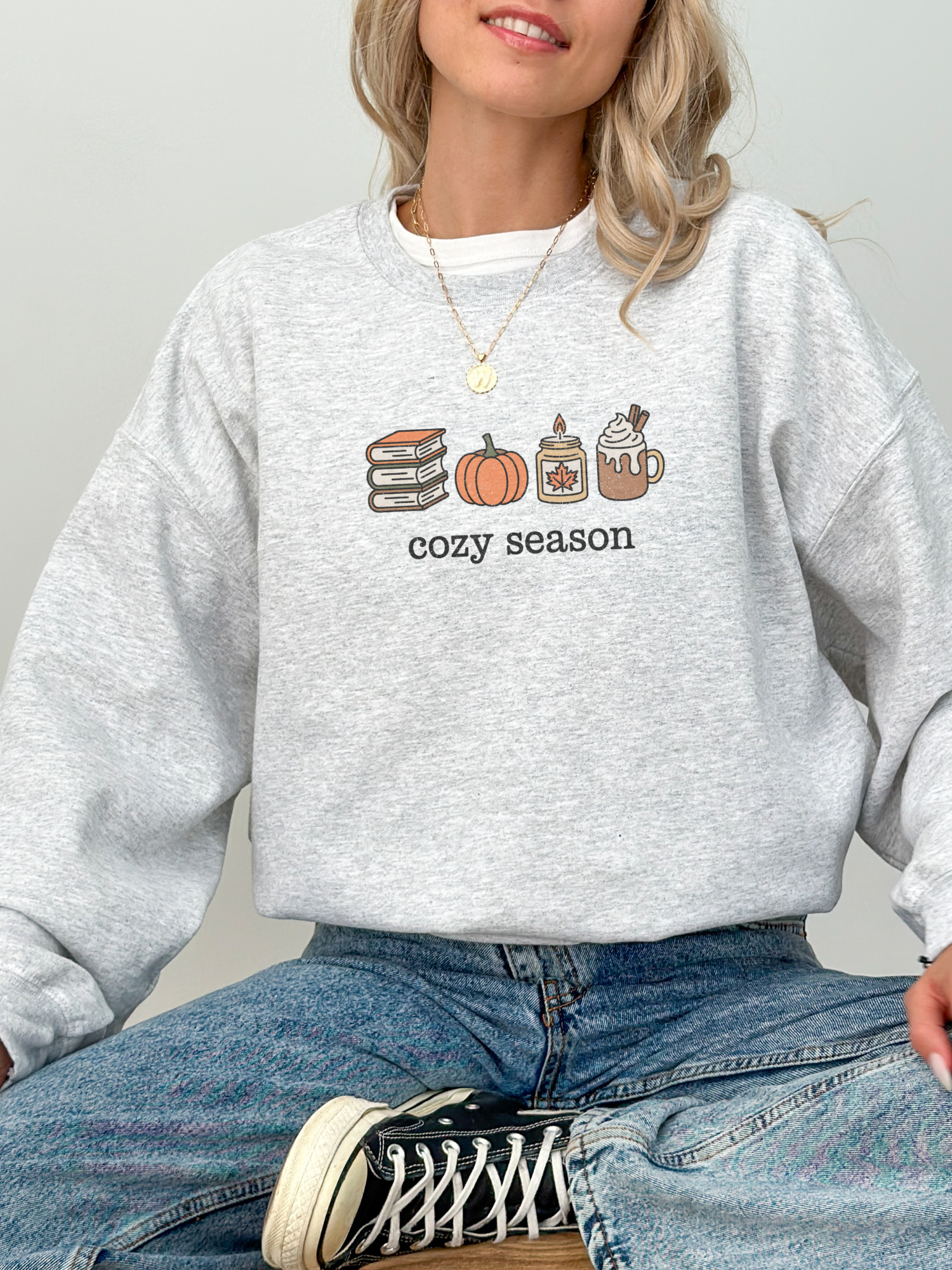 girl sitting in ash grey sweatshirt with cozy season design showing fall books, candle, pumpkin, and mug