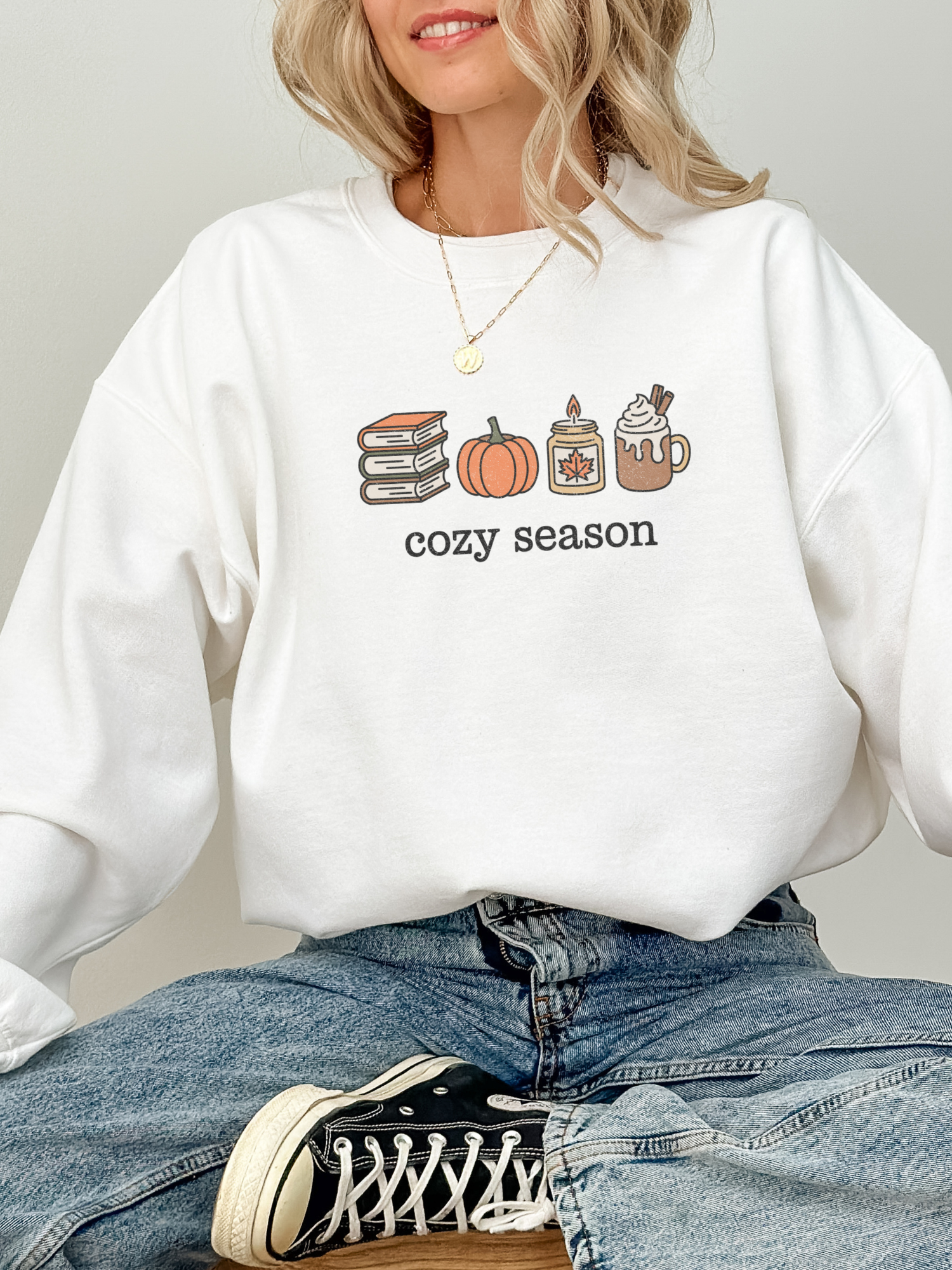 girl sitting in white sweatshirt with cozy season design featuring fall books, pumpkin, candle and hot drink