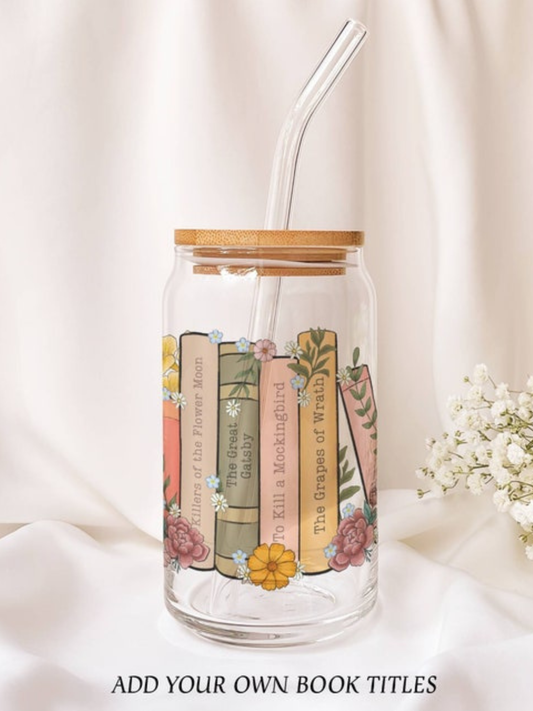 Clear glass cup with a bamboo lid and glass straw featuring a colorful stack of floral-decorated books with customizable spines, styled on a soft satin background with baby's breath.