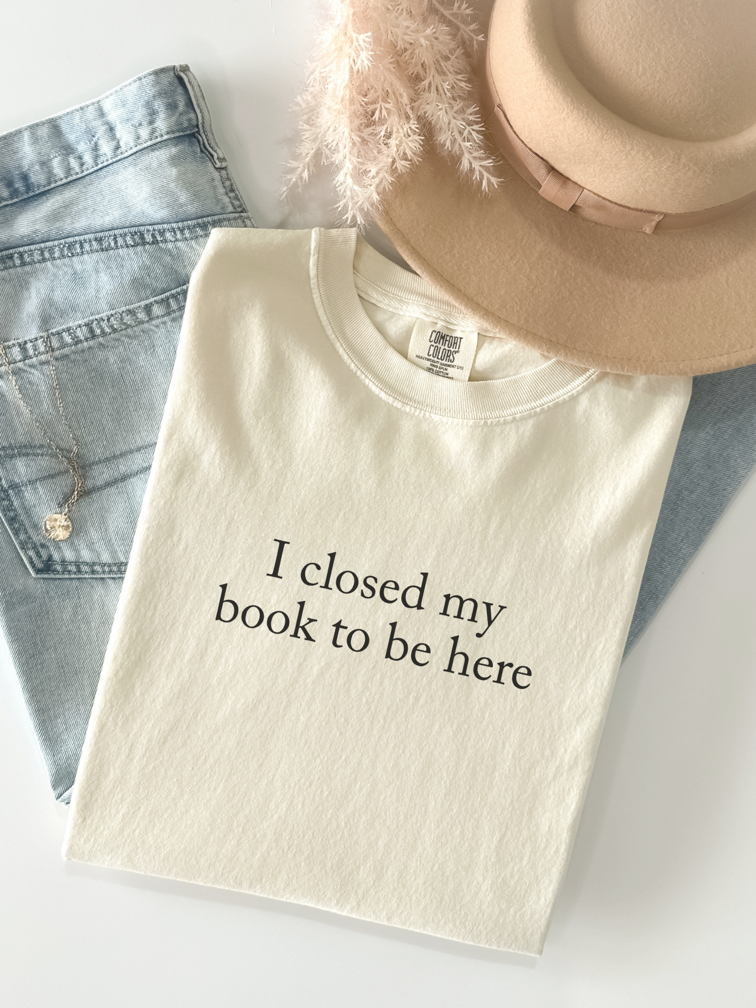 Folded ivory Comfort Colors shirt with I closed my book to be here text styled flat lay with jeans and beige hat