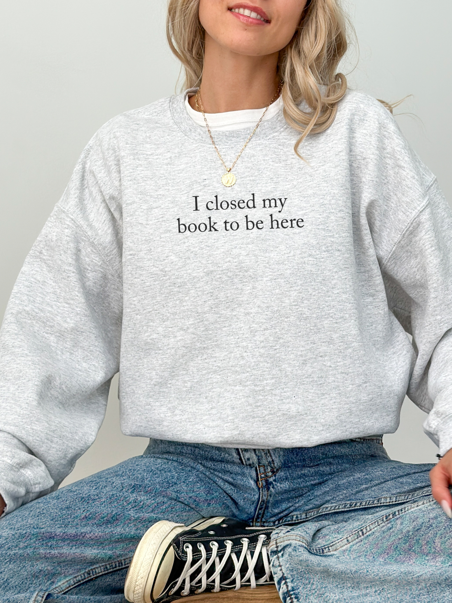 Woman sitting cross legged on a wooden stool wearing a light grey sweatshirt with the text I closed my book to be here styled with blue jeans and black high top sneakers in a minimalist studio setting