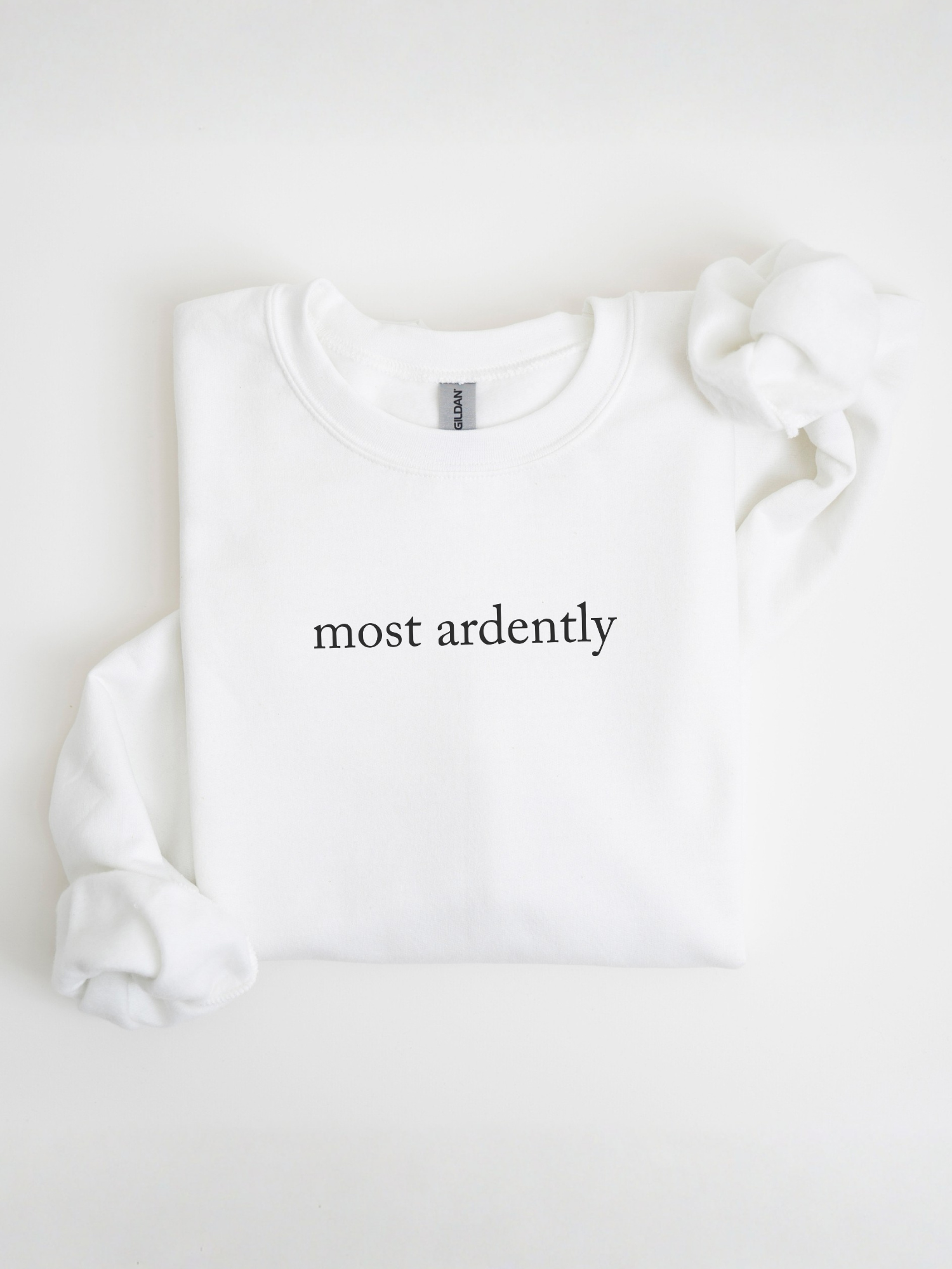 Folded white sweatshirt featuring most ardently in black text displayed in a bright flat lay setup with sleeves folded inward on a neutral background