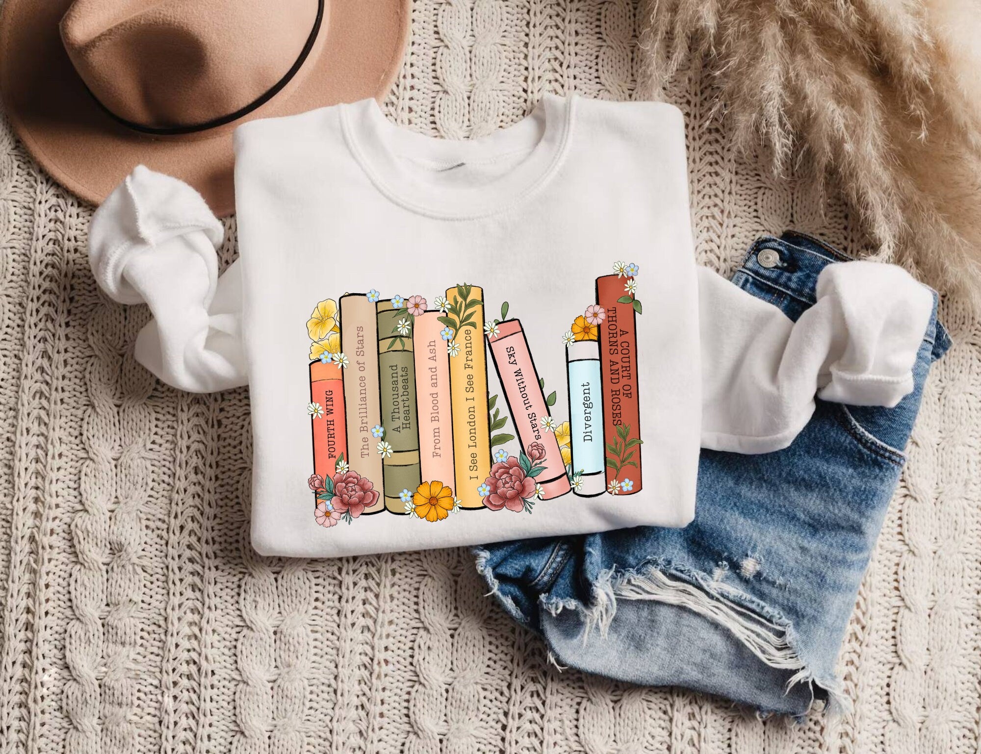 folded white sweatshirt with cozy floral bookshelf design featuring fantasy books, styled with denim shorts