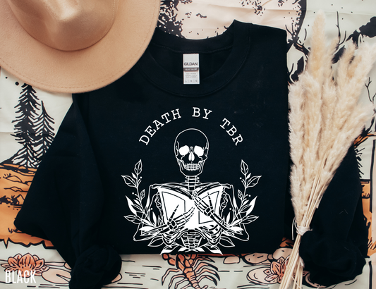 flat lay of black Death by TBR sweatshirt with white skeleton reading print styled with hat and decor