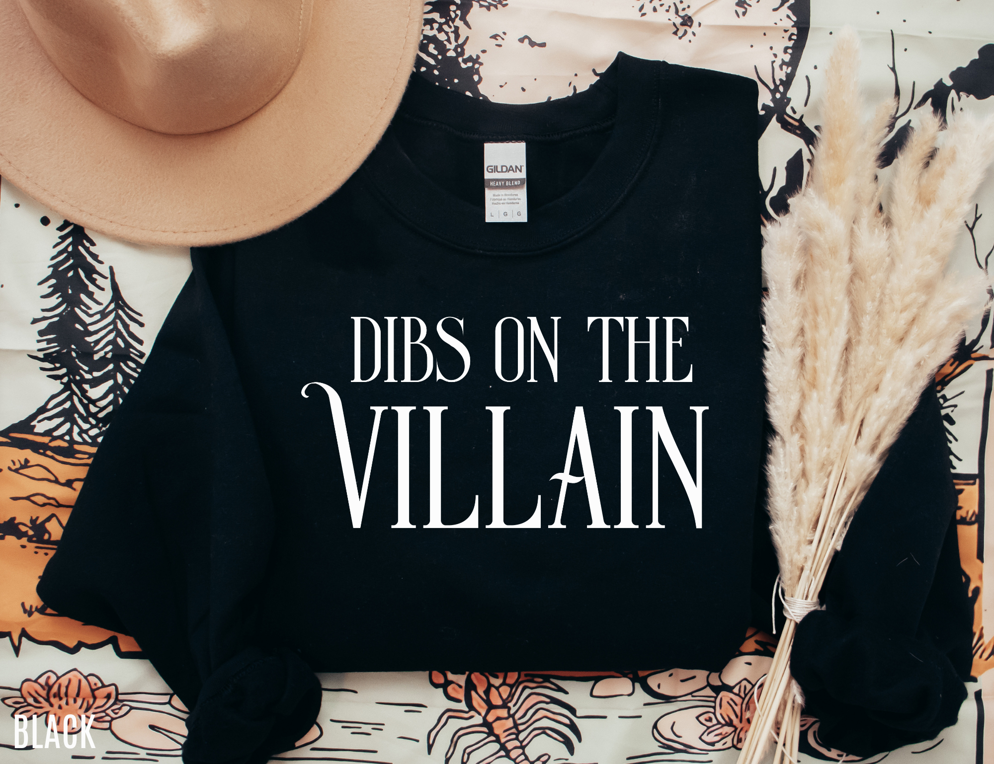 flat lay of black Dibs on the Villain sweatshirt styled with hat, book, and pampas grass