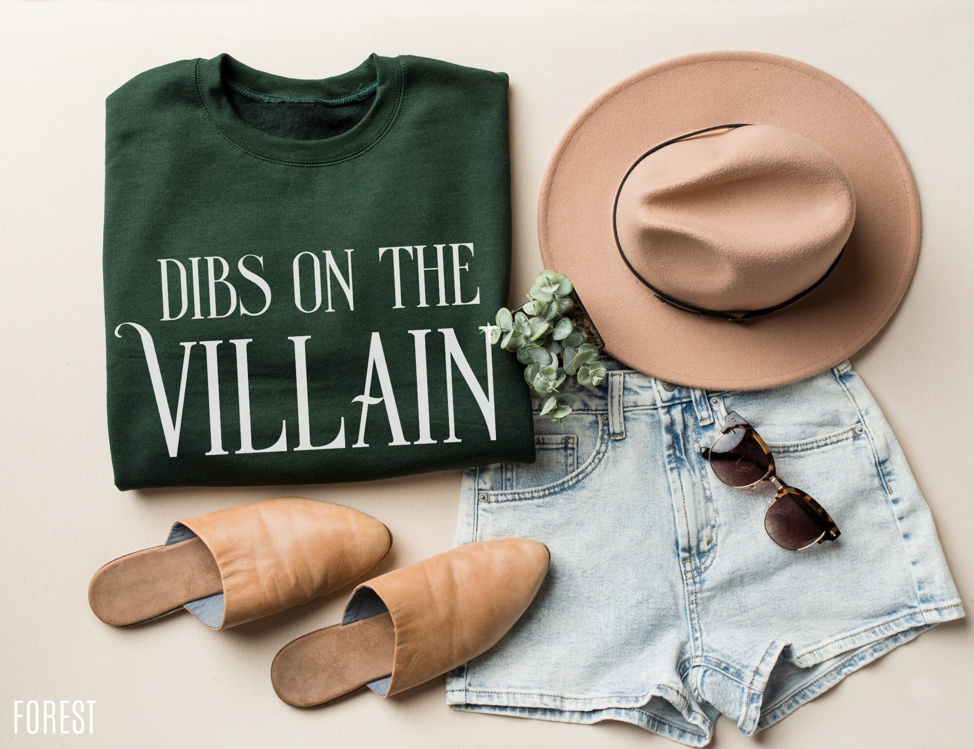 folded forest green Dibs on the Villain sweatshirt styled with sunglasses, hat, and shorts