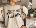 woman wearing sand-colored Dibs on the Villain sweatshirt sitting on bed in cozy room