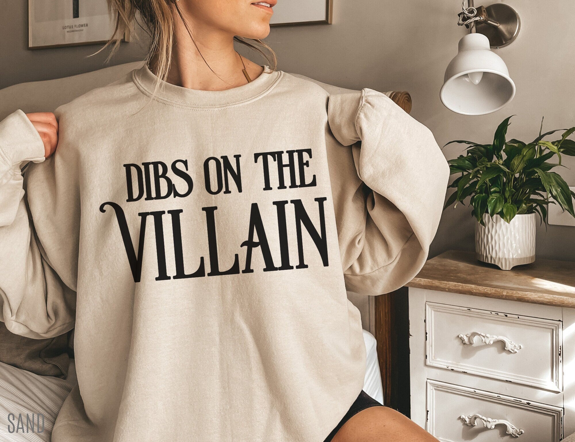 woman wearing sand-colored Dibs on the Villain sweatshirt sitting on bed in cozy room