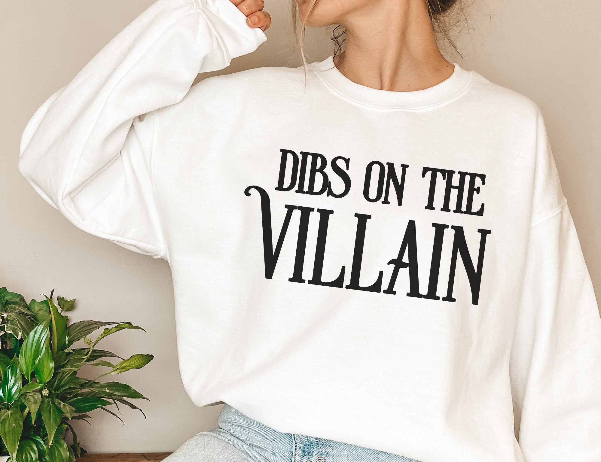 woman in oversized white sweatshirt with bold black Dibs on the Villain text, styled casually
