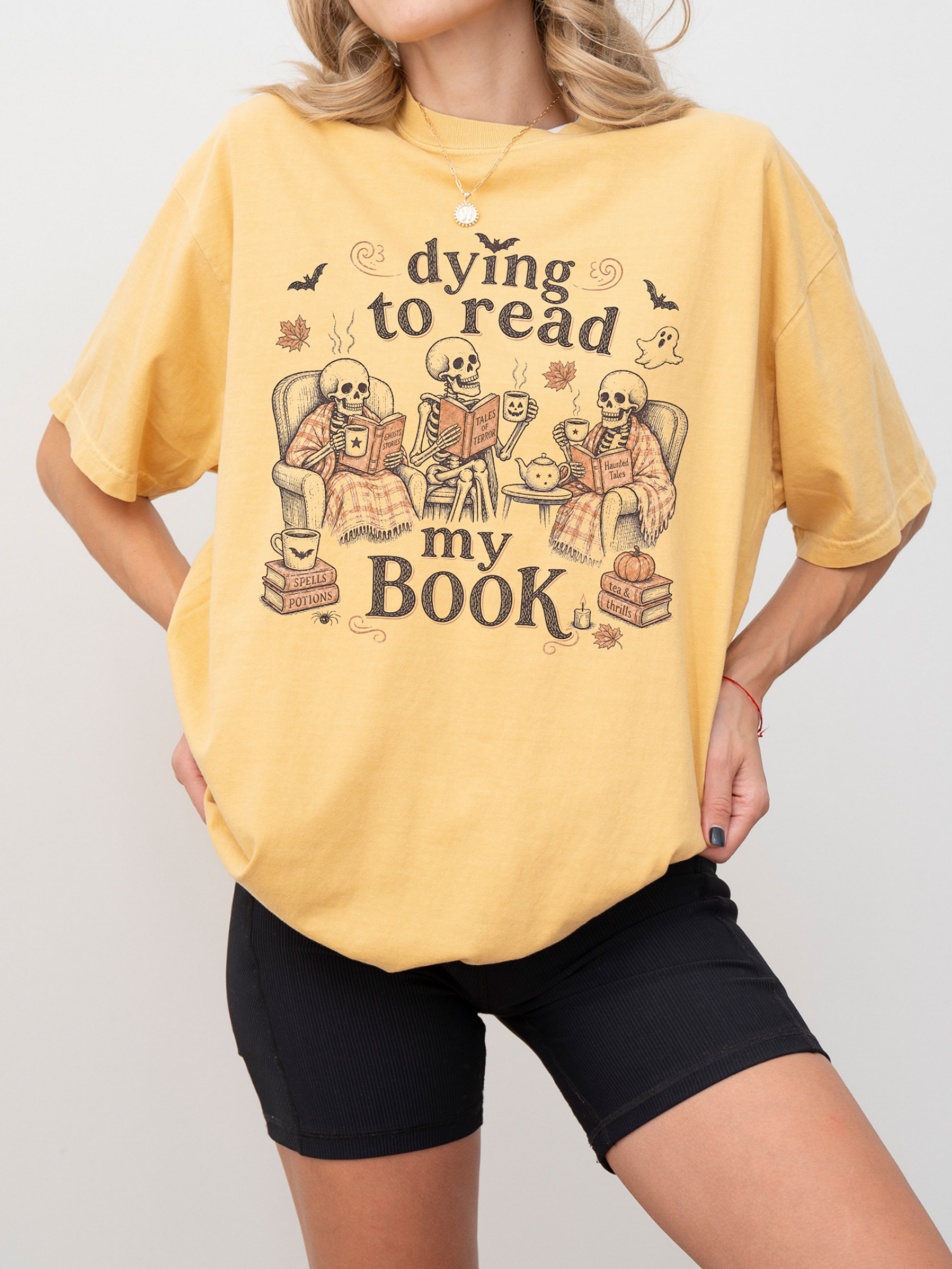 A woman wearing a mustard graphic t-shirt with a Halloween-themed skeleton illustration and the text 'dying to read my BOOK'.