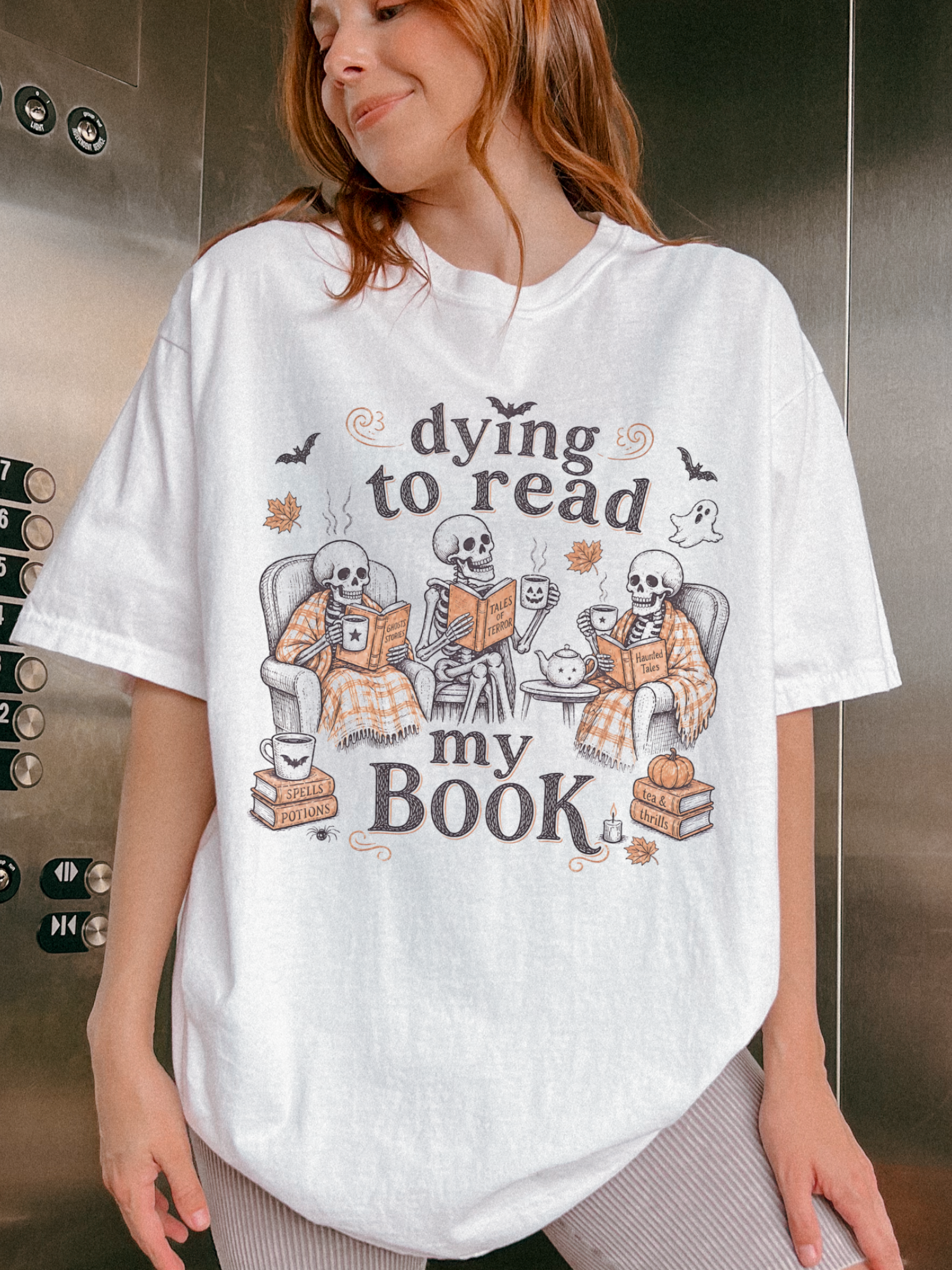 A woman wearing an oversized white graphic t-shirt with a Halloween-themed design featuring skeletons reading and the text 'dying to read my BOOK'.