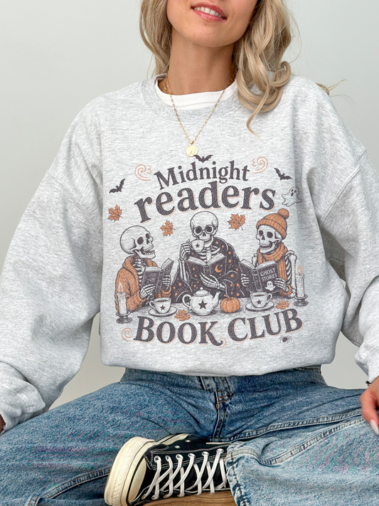 A woman wearing an ash gray crewneck sweatshirt with a Halloween-themed graphic that reads 'Midnight readers BOOK CLUB' featuring cartoon skeletons reading books. She is also wearing blue jeans and a black and white sneaker.
