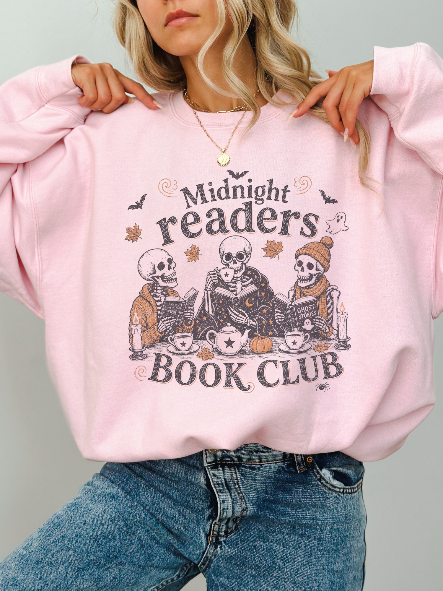A woman wearing a light pink crewneck sweatshirt with a graphic that reads 'Midnight readers BOOK CLUB' and shows three skeleton characters reading books. She is wearing blue jeans and a gold necklace against a plain light gray background.