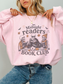 A woman wearing a light pink crewneck sweatshirt with a graphic that reads 'Midnight readers BOOK CLUB' and shows three skeleton characters reading books. She is wearing blue jeans and a gold necklace against a plain light gray background.