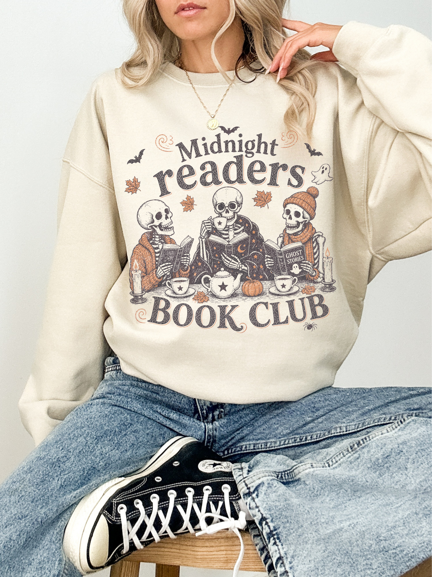 A woman wearing a sand-colored sweatshirt with a 'Midnight readers BOOK CLUB' skeleton graphic, paired with blue jeans and black sneakers.