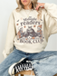 A woman wearing a sand-colored sweatshirt with a 'Midnight readers BOOK CLUB' skeleton graphic, paired with blue jeans and black sneakers.