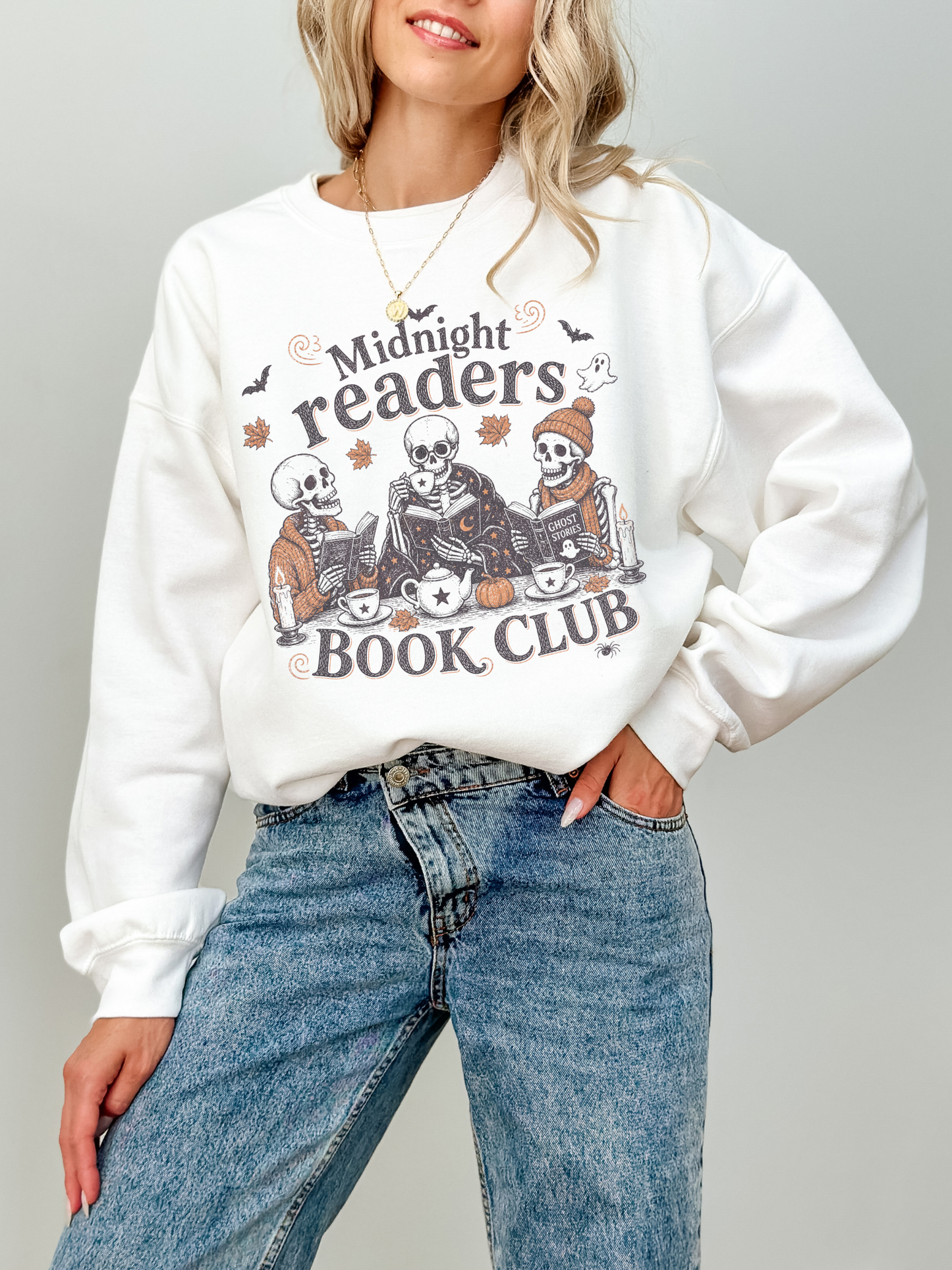 A woman wearing a white crewneck sweatshirt with a Halloween-themed graphic that reads 'Midnight readers BOOK CLUB' featuring cartoon skeletons reading, paired with blue jeans, against a solid light gray background.