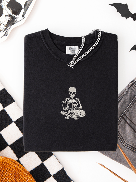 A folded black t-shirt with a white skeleton embroidered graphic sitting cross-legged reading, styled in a flat lay with a silver chain necklace, a checkered blanket, and Halloween props on a white background.