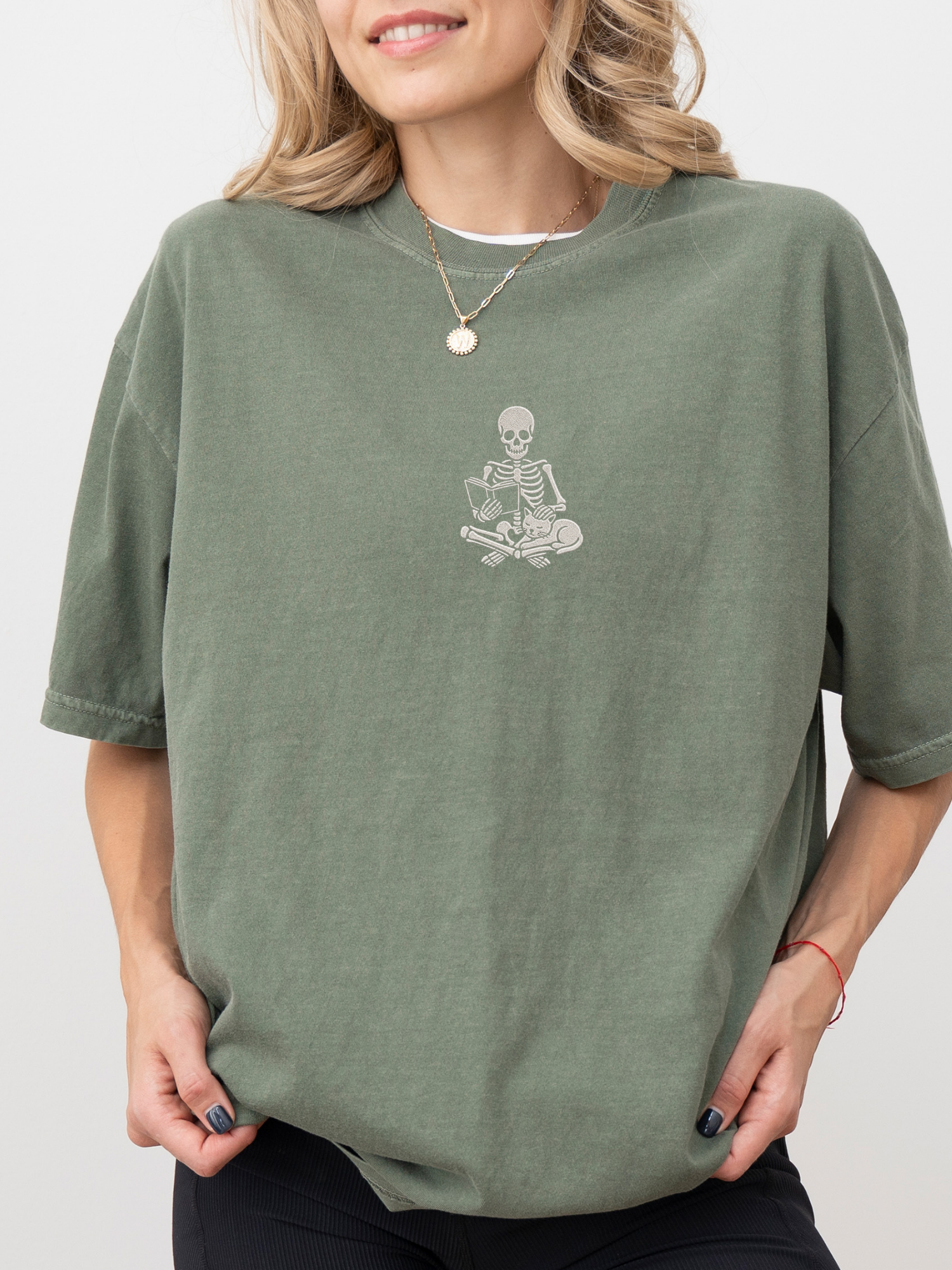 A woman wearing an oversized moss green t-shirt with a small white embroidered graphic of a skeleton meditating on the chest, set against a plain light gray background.