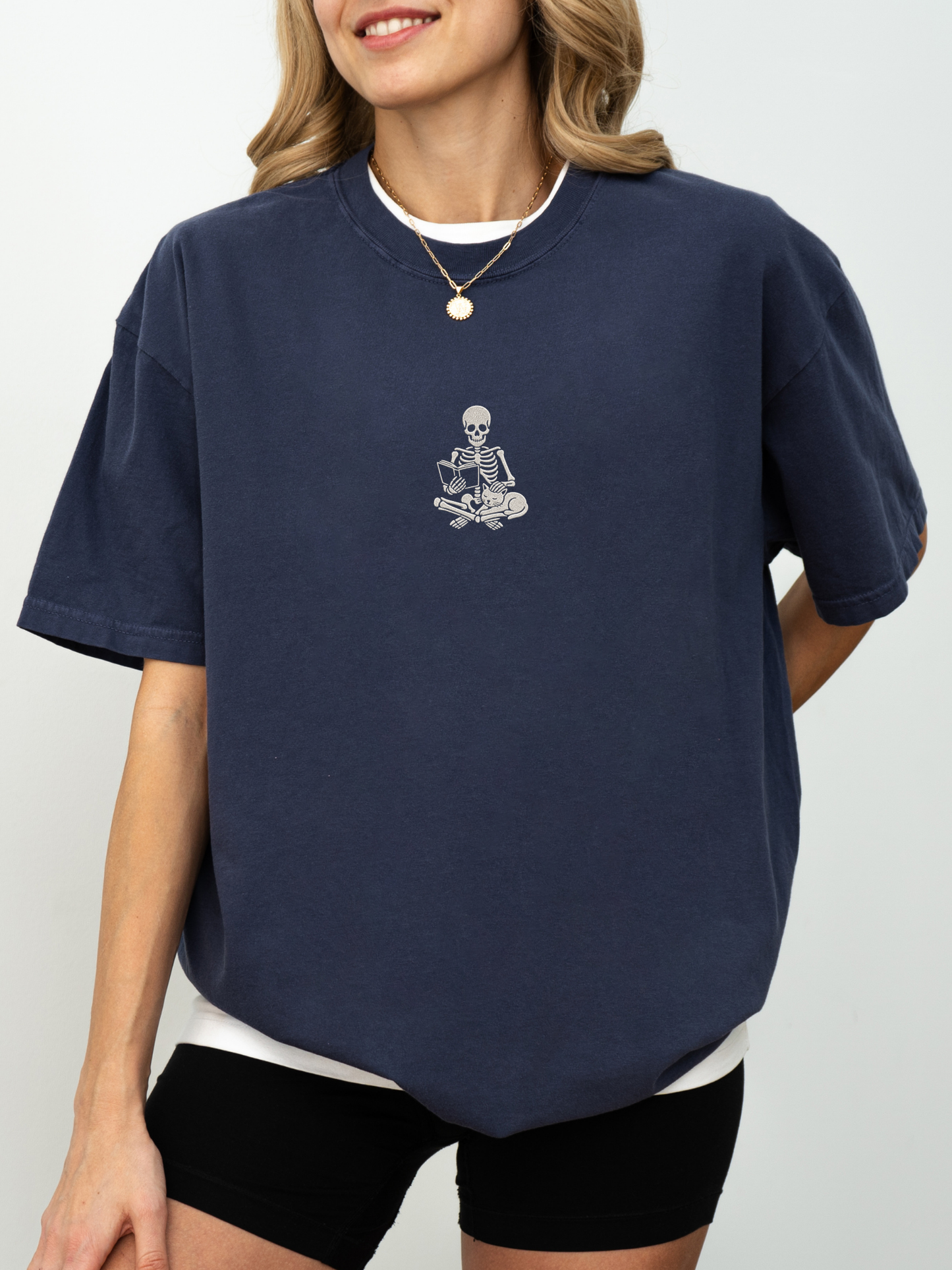 A woman wearing an oversized navy blue t-shirt with a small embroidered white graphic of a skeleton sitting on a bicycle on the left chest, paired with black bike shorts and a gold necklace.