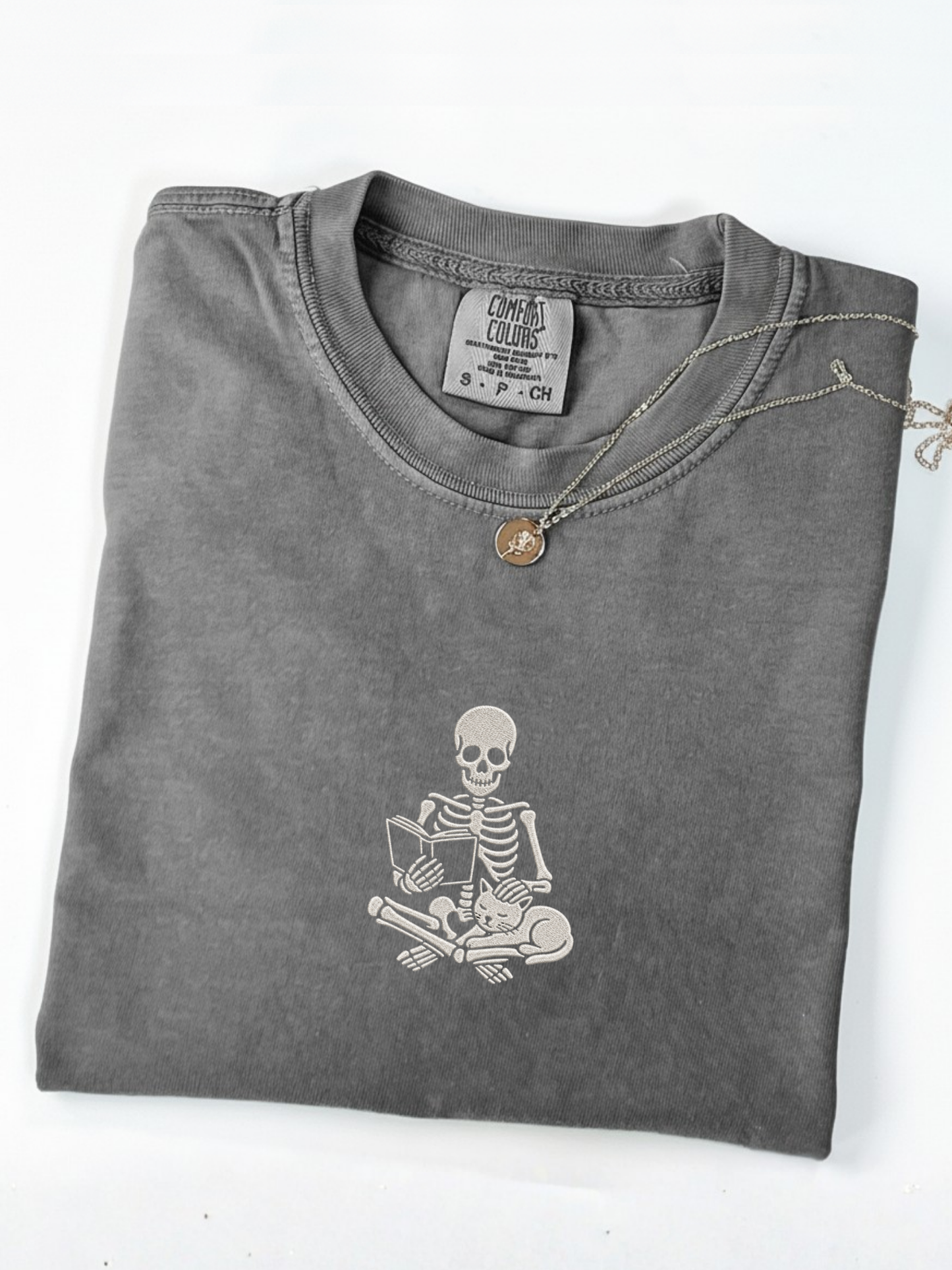 A folded pepper t-shirt with an embroidered white graphic of a skeleton reading a book on the front, styled with a silver chain necklace featuring a small round pendant, on a white background.