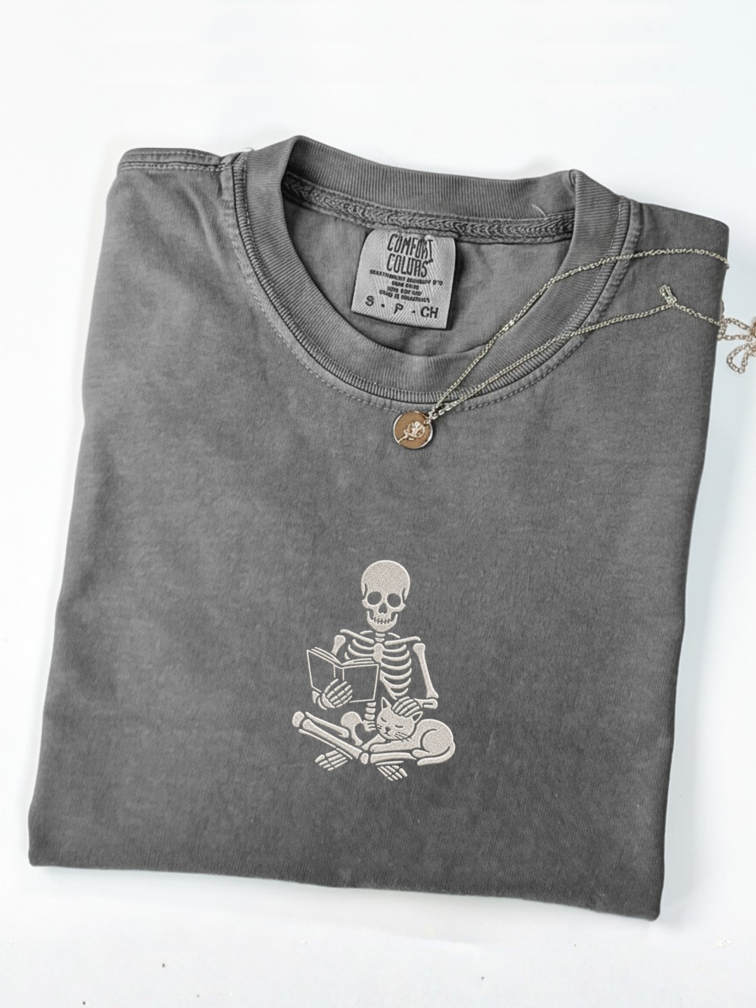 A folded pepper t-shirt with an embroidered white graphic of a skeleton reading a book on the front, styled with a silver chain necklace featuring a small round pendant, on a white background.
