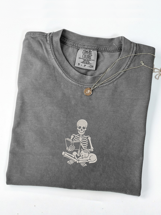 A folded pepper t-shirt with an embroidered white graphic of a skeleton reading a book on the front, styled with a silver chain necklace featuring a small round pendant, on a white background.