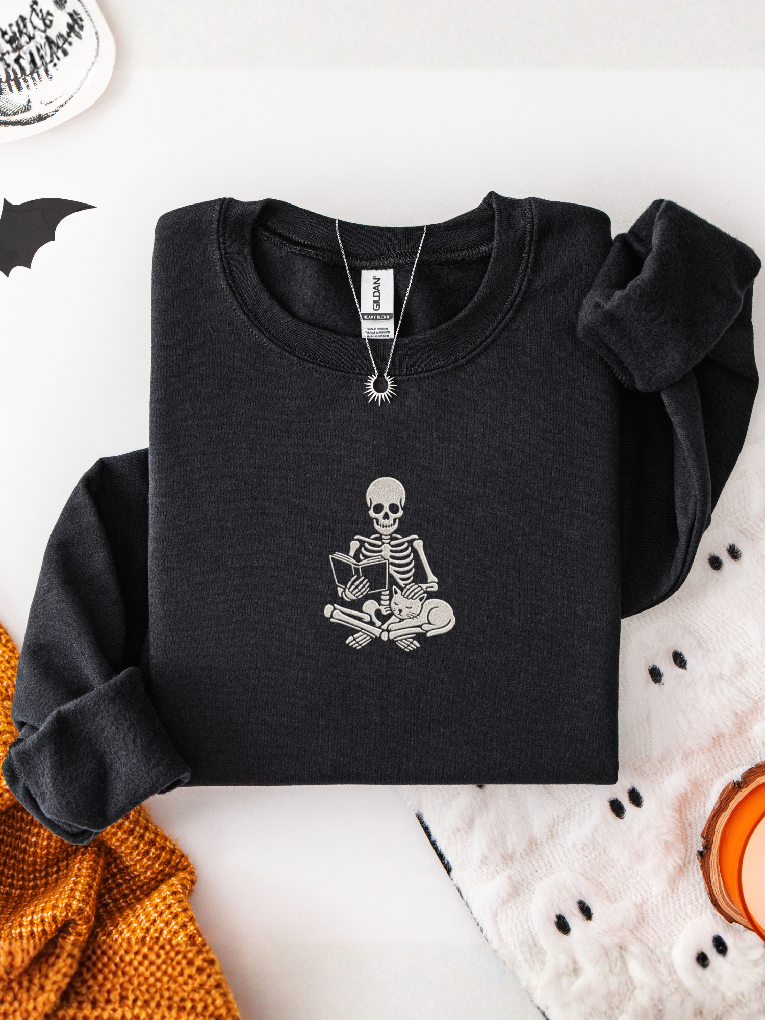 A folded black long-sleeve sweatshirt with a white embroidered skeleton graphic sitting cross-legged on the chest, styled on a white surface with Halloween-themed props including a knit orange sweater, a white knit blanket, and a necklace.