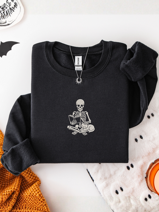 A folded black long-sleeve sweatshirt with a white embroidered skeleton graphic sitting cross-legged on the chest, styled on a white surface with Halloween-themed props including a knit orange sweater, a white knit blanket, and a necklace.