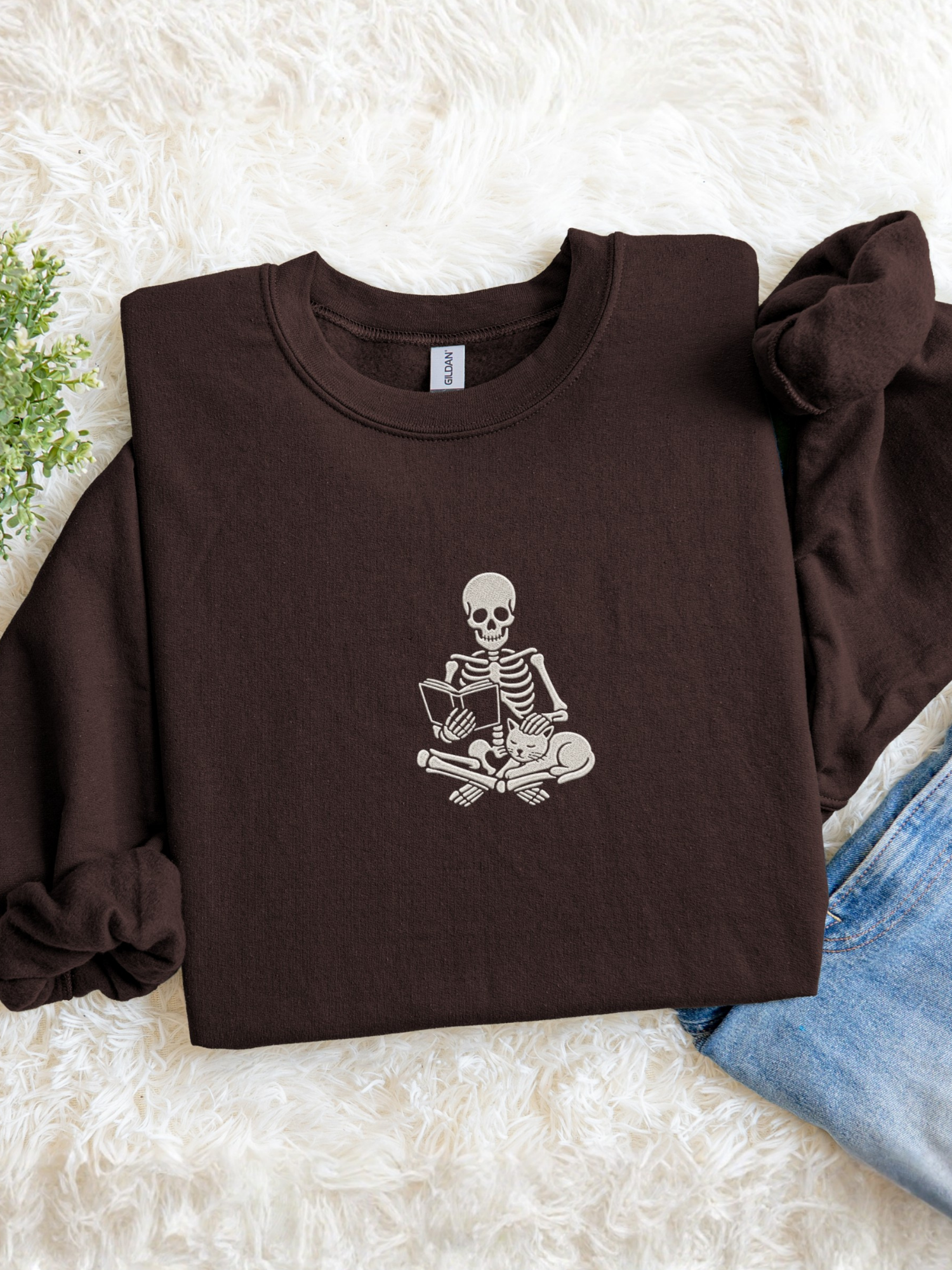 A dark chocolate crewneck sweatshirt with a white embroidered graphic of a skeleton sitting cross-legged reading a book, styled in a flat lay on a white faux fur rug with blue jeans and a small green plant.