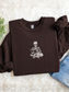 A dark chocolate crewneck sweatshirt with a white embroidered graphic of a skeleton sitting cross-legged reading a book, styled in a flat lay on a white faux fur rug with blue jeans and a small green plant.