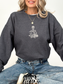 A woman wearing a dark heather gray crewneck sweatshirt with a white embroidered graphic of a skeleton sitting cross-legged on the chest, paired with blue jeans and a black-and-white sneaker.