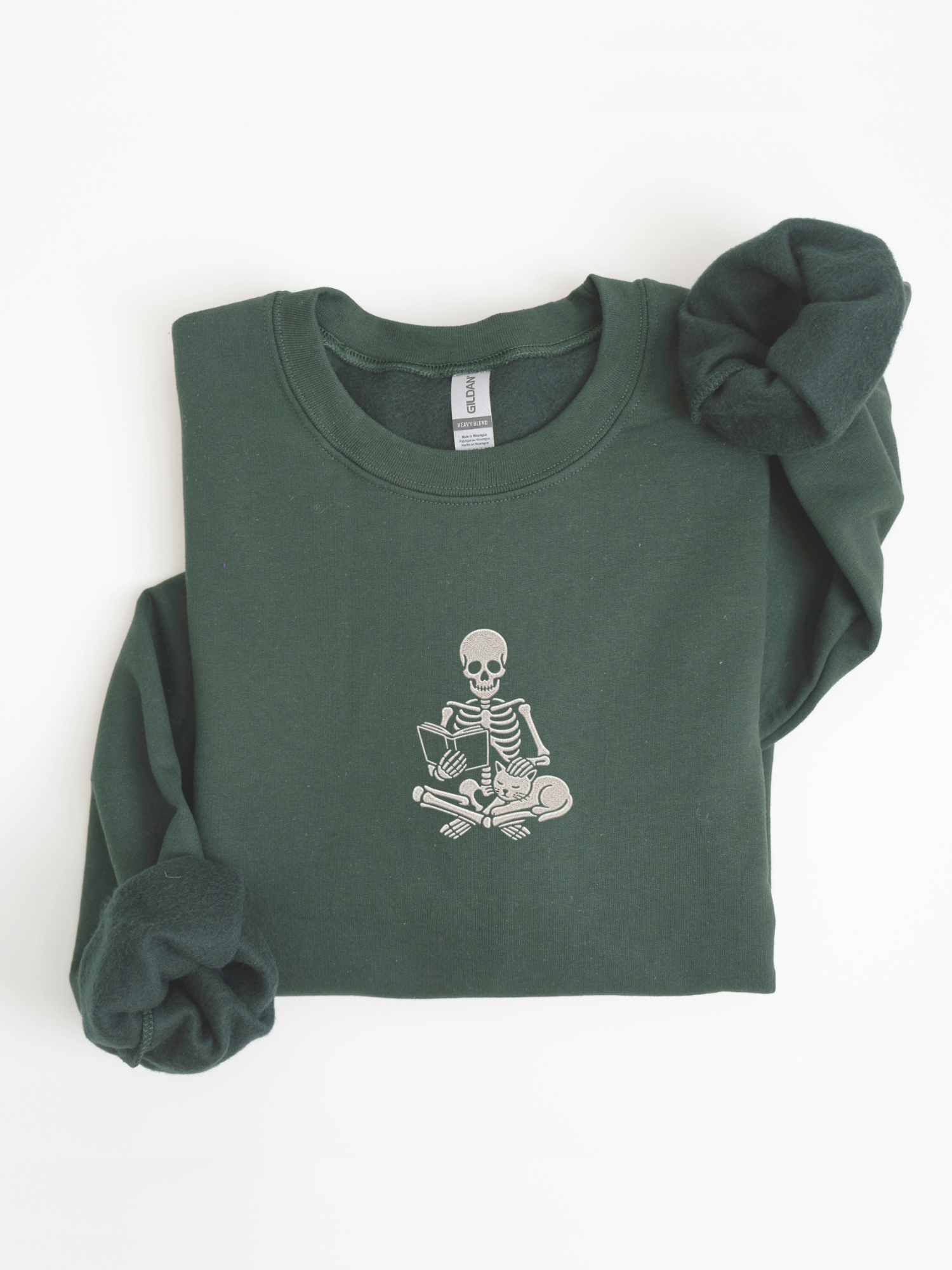 A forest green long-sleeve sweatshirt with a white embroidered graphic of a skeleton sitting cross-legged reading a book on the chest, shown folded on a white background.