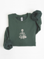 A forest green long-sleeve sweatshirt with a white embroidered graphic of a skeleton sitting cross-legged reading a book on the chest, shown folded on a white background.
