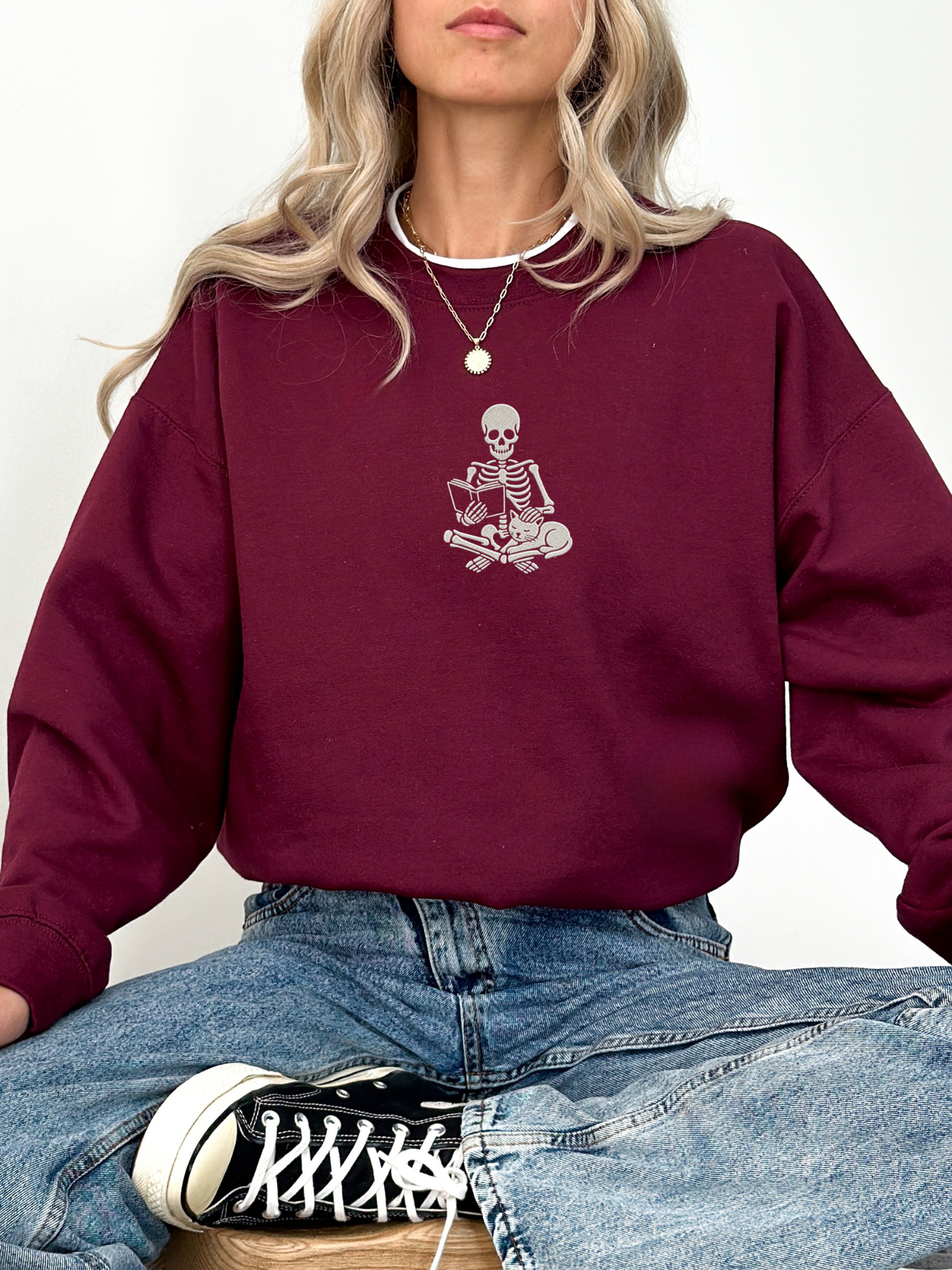 A woman wearing a maroon crewneck sweatshirt with a white embroidered skeleton graphic sitting on crossed bones on the chest, paired with blue jeans and a black and white canvas sneaker.