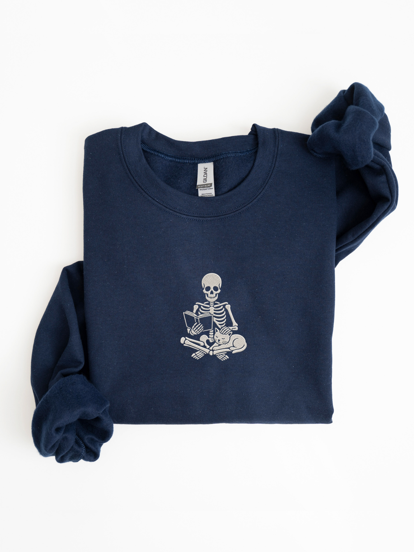 A folded navy blue sweatshirt with a white embroidered graphic of a skeleton sitting cross-legged reading a book, displayed on a clean white background.