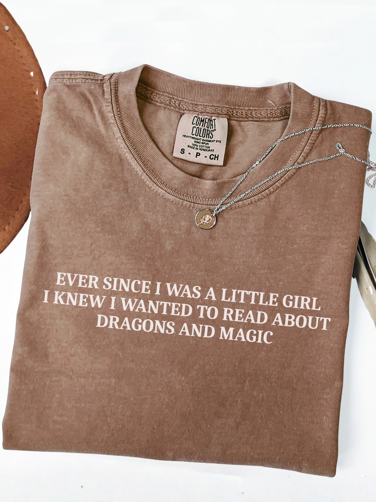 A folded espresso brown t-shirt with white text that reads 'EVER SINCE I WAS A LITTLE GIRL I KNEW I WANTED TO READ ABOUT DRAGONS AND MAGIC'. A silver necklace with a gold charm is draped over the collar.