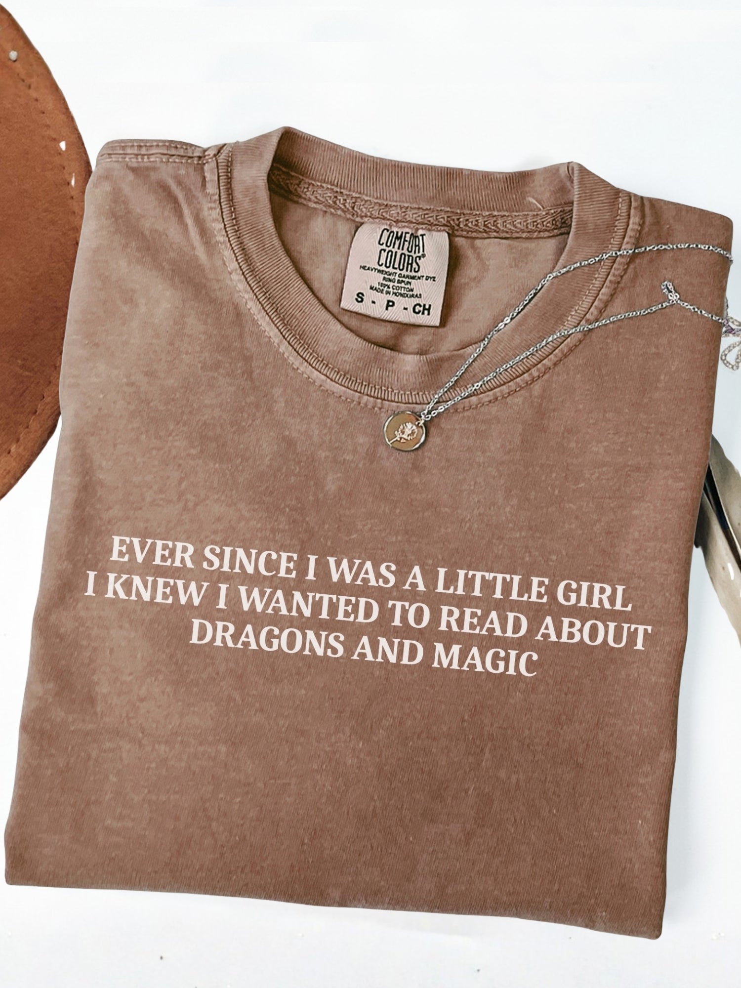 A folded espresso brown t-shirt with white text that reads 'EVER SINCE I WAS A LITTLE GIRL I KNEW I WANTED TO READ ABOUT DRAGONS AND MAGIC'. A silver necklace with a gold charm is draped over the collar.