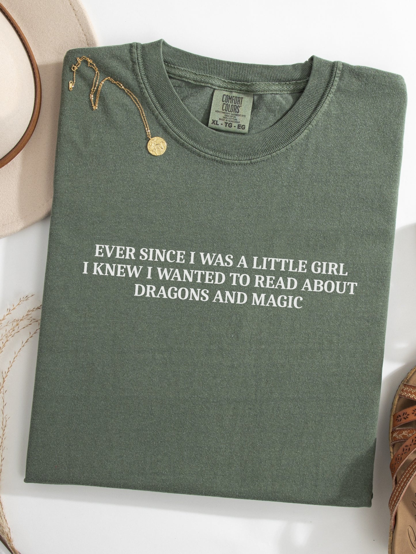 A moss green folded t-shirt with white printed text reading 'EVER SINCE I WAS A LITTLE GIRL I KNEW I WANTED TO READ ABOUT DRAGONS AND MAGIC', styled on a white surface with a gold necklace and a beige hat partially visible.