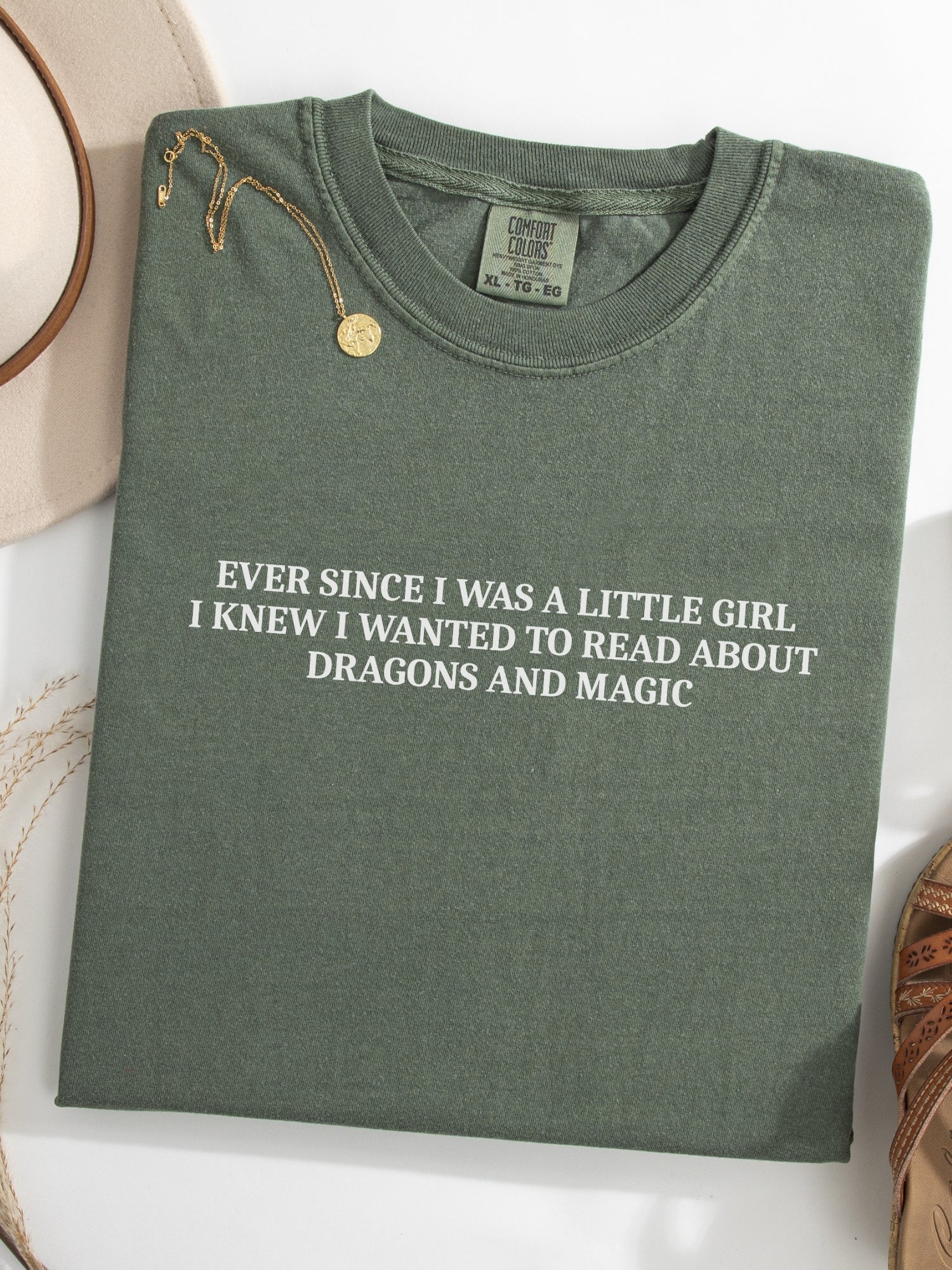 A moss green folded t-shirt with white printed text reading 'EVER SINCE I WAS A LITTLE GIRL I KNEW I WANTED TO READ ABOUT DRAGONS AND MAGIC', styled on a white surface with a gold necklace and a beige hat partially visible.